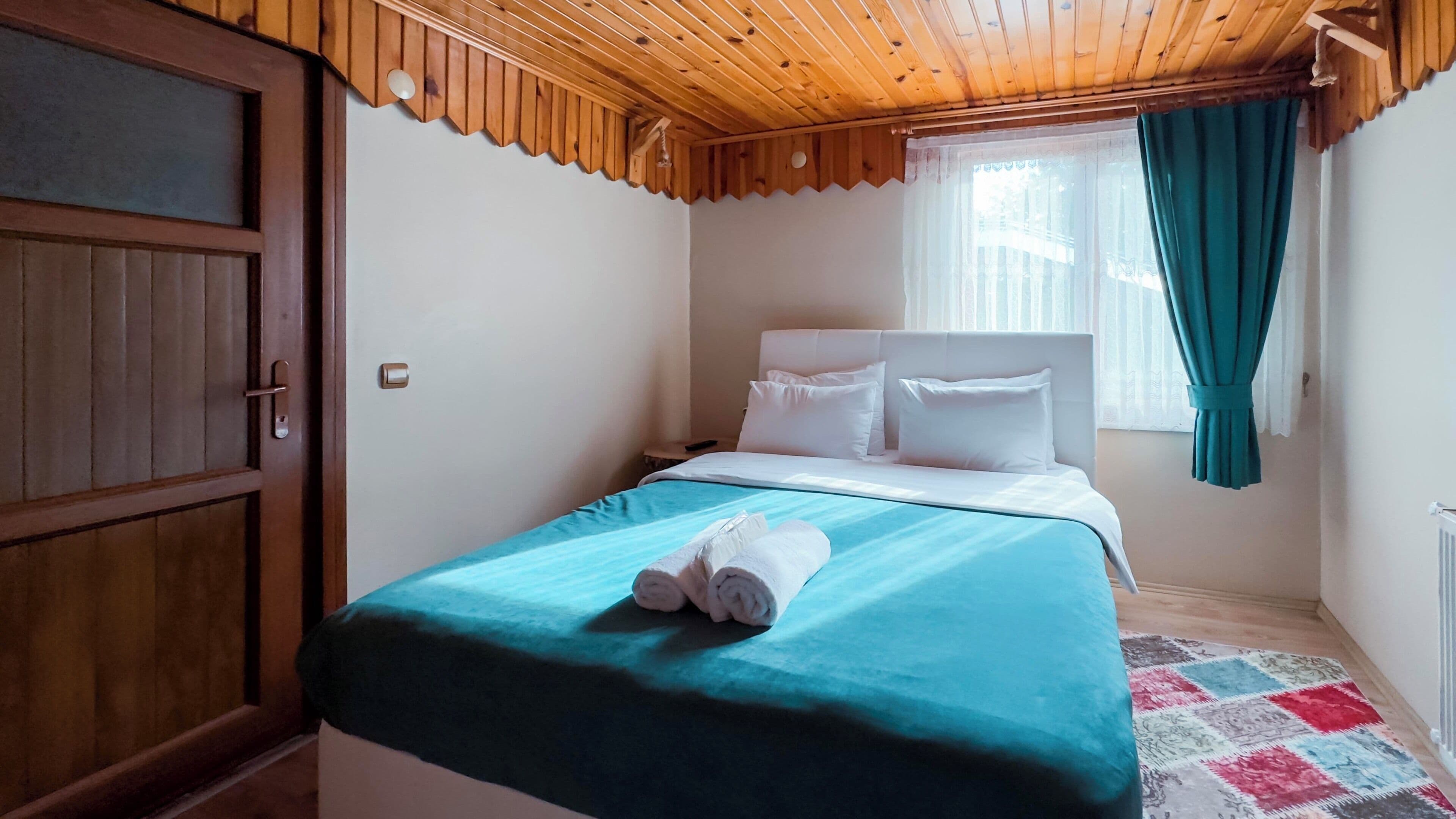 A bedroom with a wooden ceiling, a bed with white bedding, a teal bedspread, and a rolled towel on the bed.