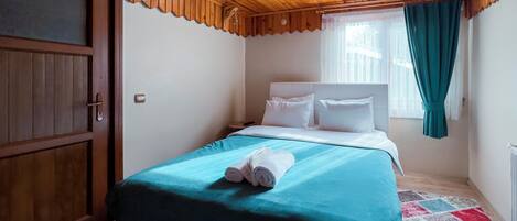 A bedroom with a wooden ceiling, a bed with white bedding, a teal bedspread, and a rolled towel on the bed.