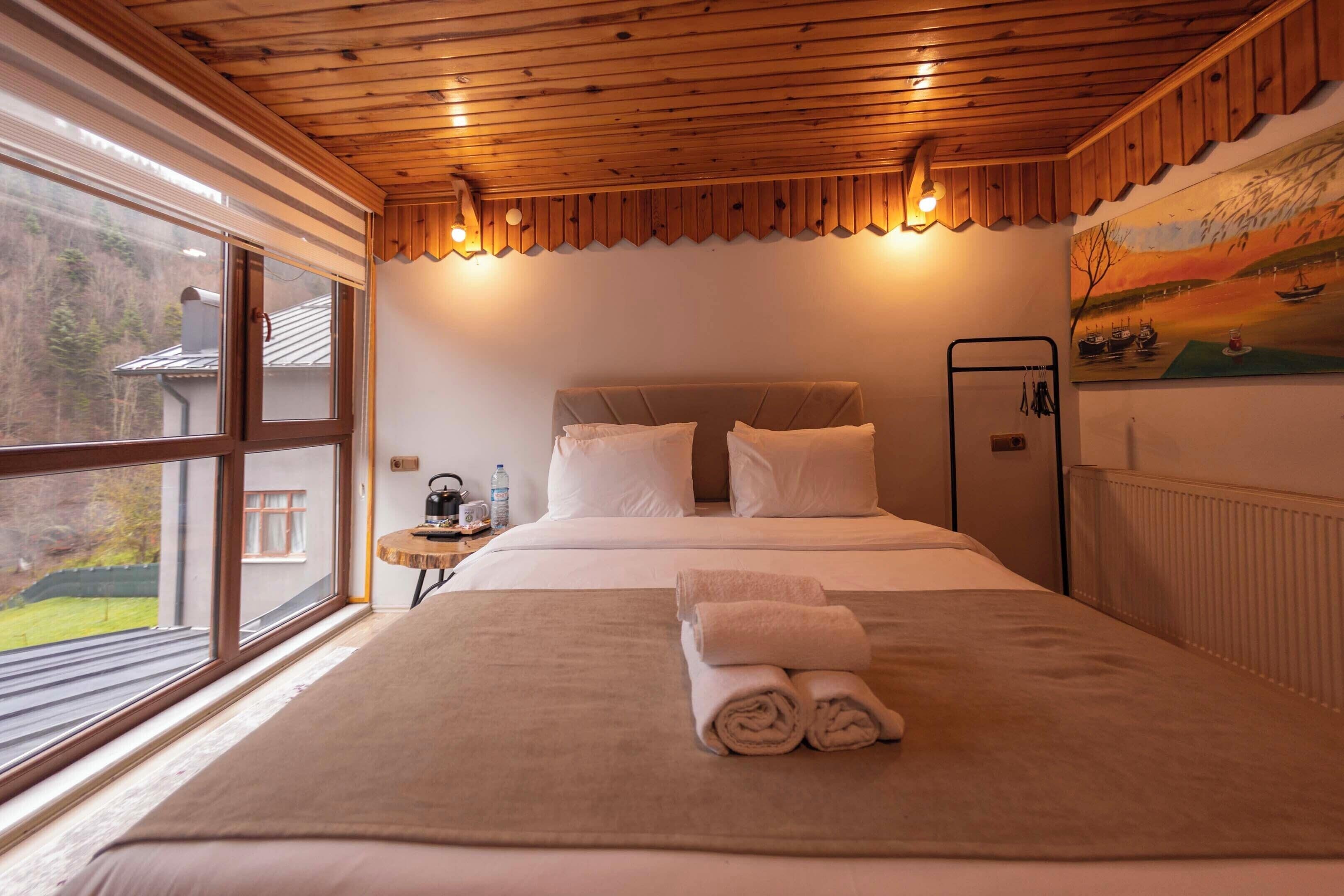 A bedroom with a large bed, a wooden ceiling, a painting on the wall, and a window with a view of the outside.