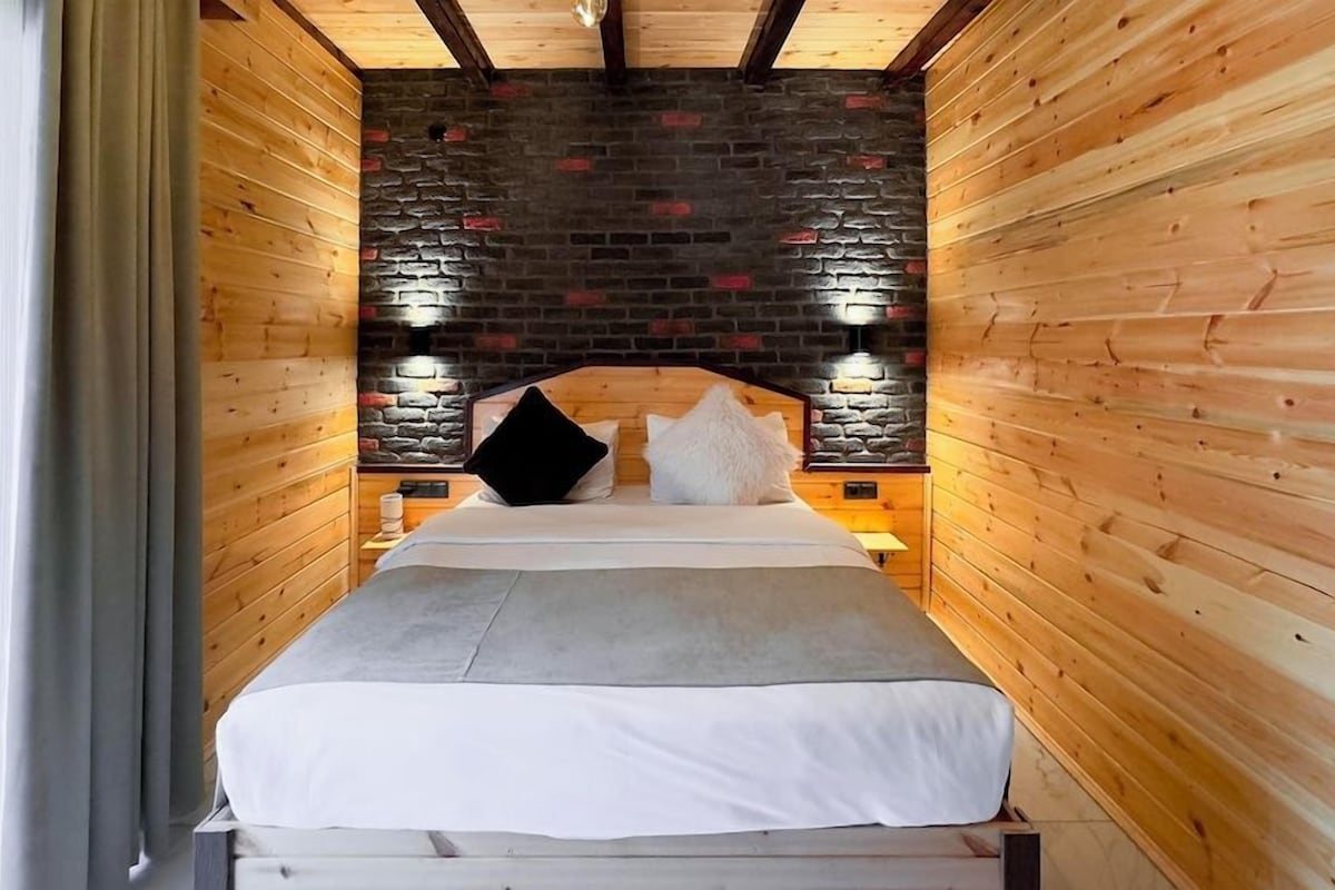 A narrow, wooden-paneled room with a bed centered against a brick wall, flanked by two wall-mounted lights.