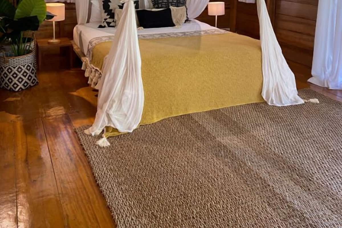 A room with a four-poster bed, a wooden floor, a large potted plant, and a window with curtains.