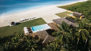 Vue aérienne d’un complexe hôtelier de bord de mer, avec une piscine, des chaises longues et des cabanes à toit de chaume.