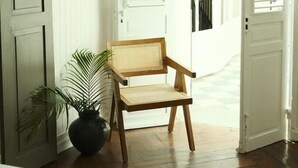A wooden chair with a woven backrest placed in a hallway with white doors and a potted plant. - Maison Victoire (Pointe-à-Pitre)