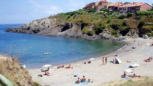 A beach with people sunbathing and swimming, a rocky shoreline, and buildings on a hill in the background. - T2 Large terrace with sea view & direct access to the beach – Collioure sector (Port-Vendres)