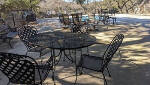 Un patio con mesas y sillas de metal, piscina y un área cercada con árboles.