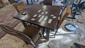 Una mesa de madera con cartas de juego colocadas encima, rodeada de sillas tapizadas con un diseño estampado.