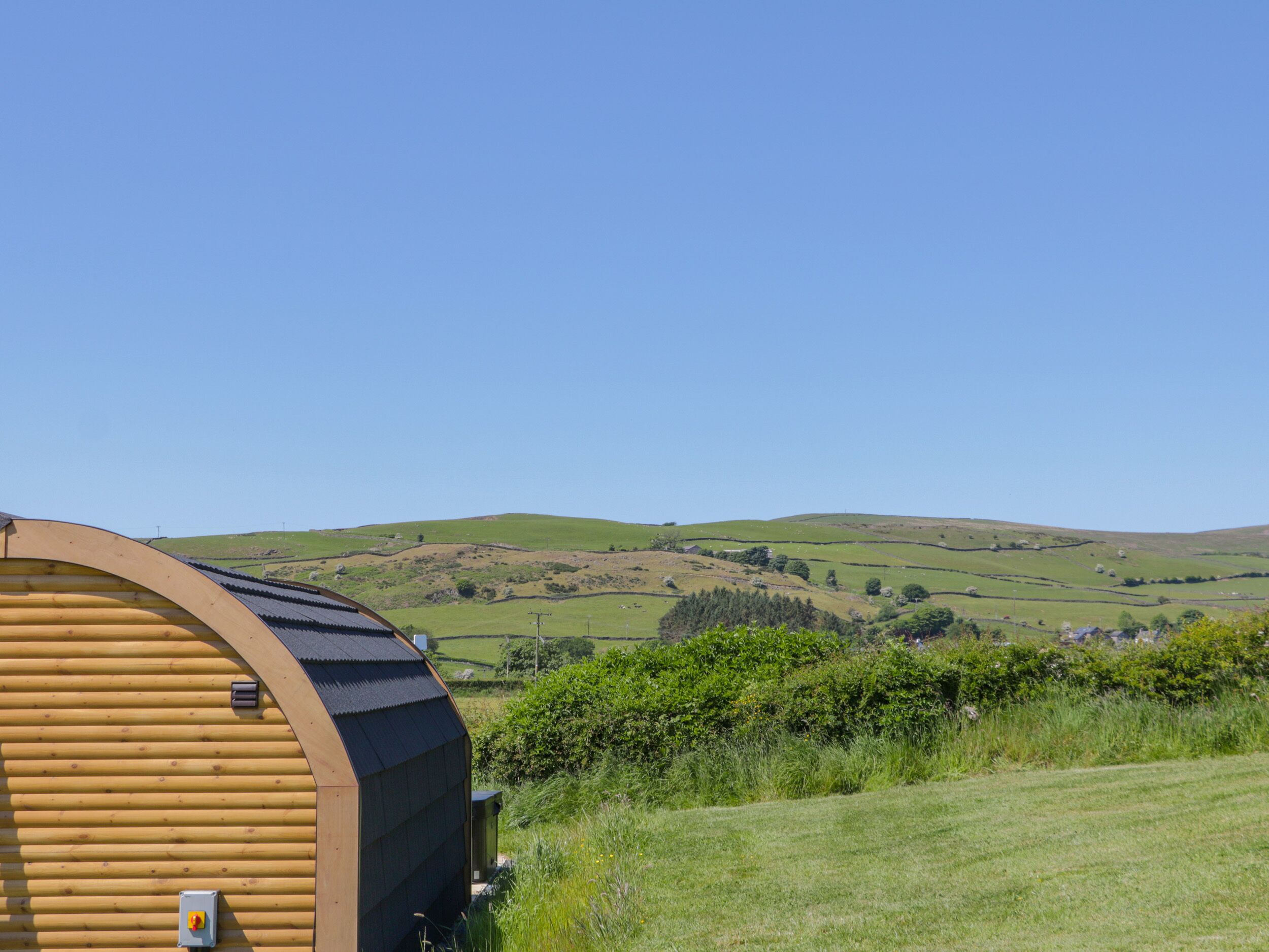 A wooden cabin with a curved roof, surrounded by green hills and a clear sky.