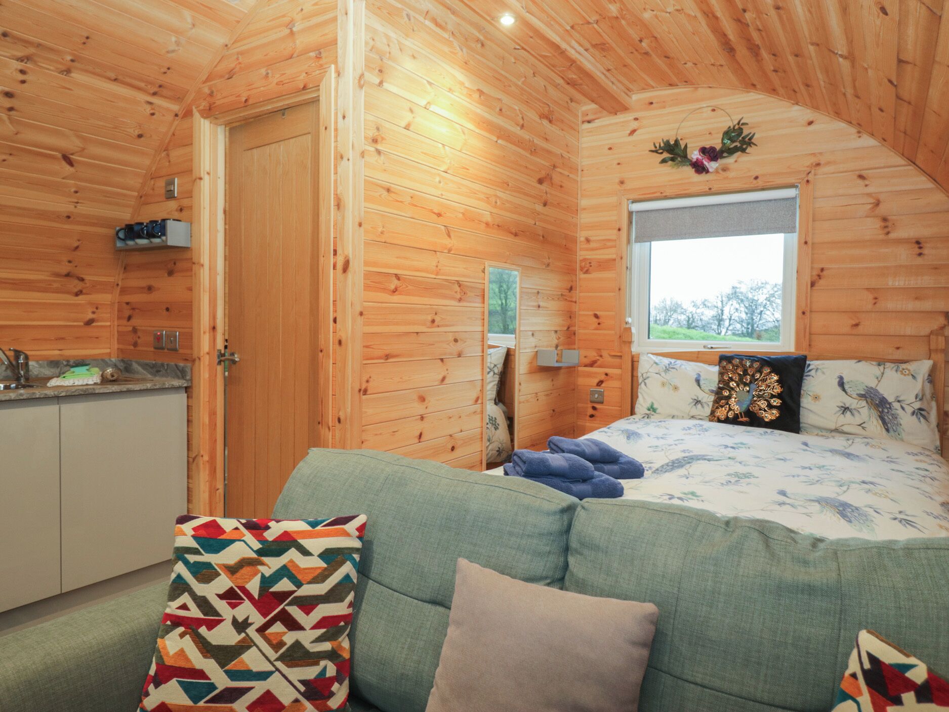 A cozy cabin interior with a wooden interior, a bed with floral bedding, a green sofa, and a window with a view of trees.