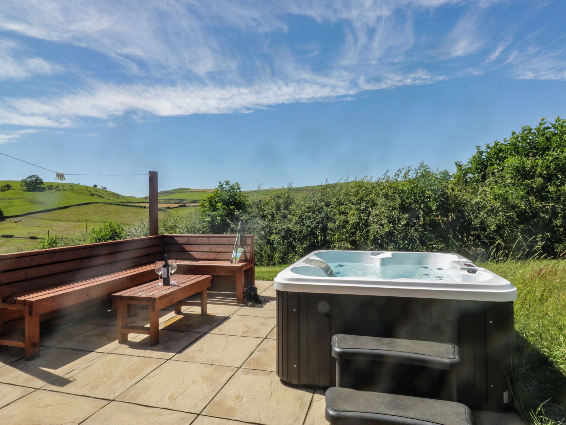 A hot tub with a wooden bench and a table, set on a patio with a view of green hills.
