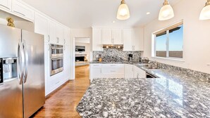 Une cuisine moderne avec des armoires blanches, des appareils en acier inoxydable, des comptoirs en granit et des luminaires suspendus.