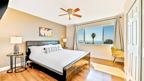 Une chambre moderne avec un grand lit, un ventilateur de plafond, une vue sur l’océan et un fauteuil près de la fenêtre.