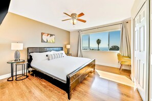 Ein modernes Schlafzimmer mit einem großen Bett, einem Deckenventilator, Blick auf den Ozean und einem Sessel am Fenster.