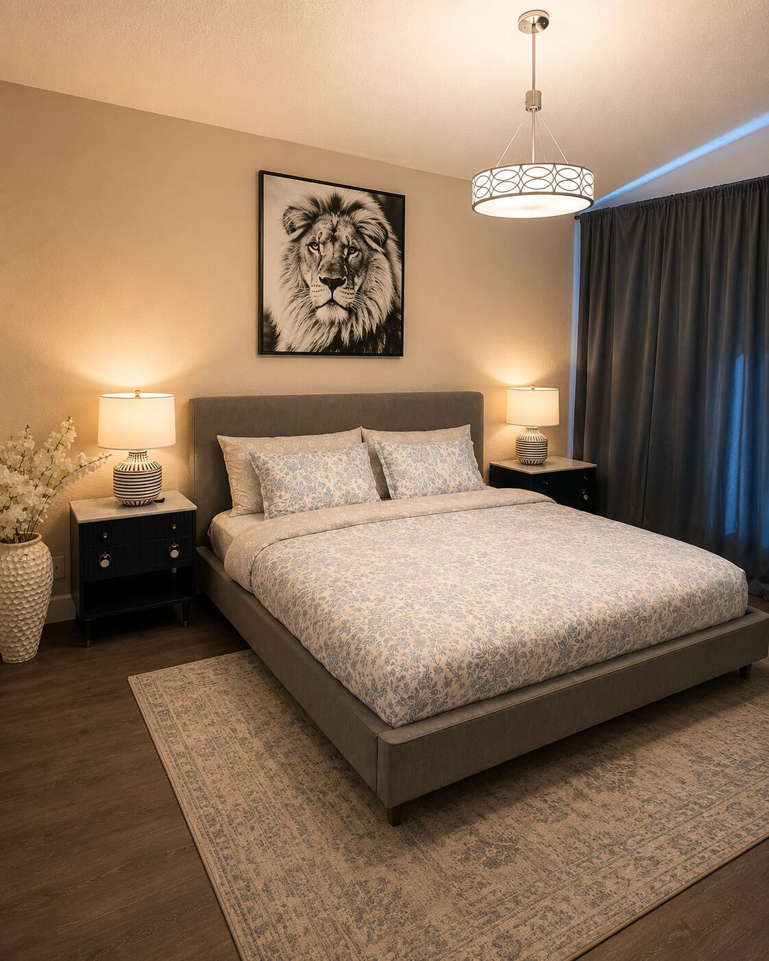 A bedroom with a large bed, two bedside tables, a wall-mounted lamp, a framed picture, and a window with curtains.
