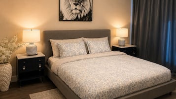 A bedroom with a large bed, two bedside tables, a wall-mounted lamp, a framed picture, and a window with curtains.