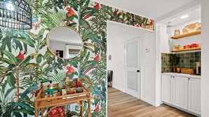 Um quarto com papel de parede com tema tropical, um espelho redondo, uma mesinha de vime com diversos objetos, e uma área de cozinha com armários brancos e painel de azulejos.