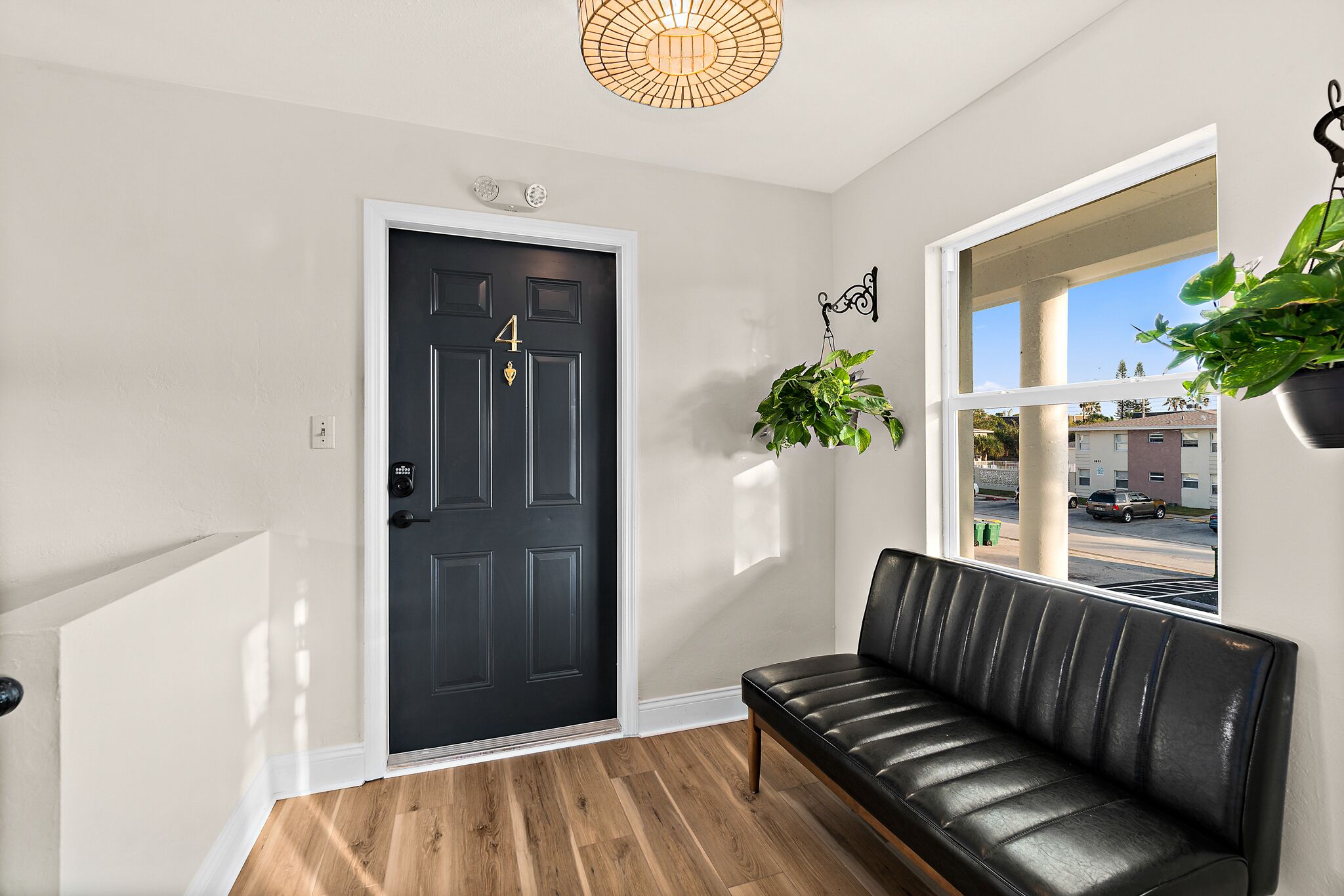 A black door with the number 4, a black leather bench, a hanging plant, and a window with a view of buildings and a street.
