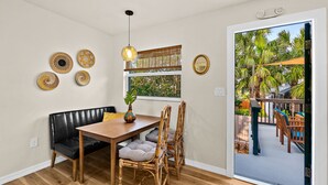 Uma área de refeições com uma mesa de madeira, um banco de couro preto e cadeiras de bambu. Há decorações nas paredes e uma janela com vista para palmeiras.