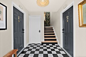 Ein Korridor mit schwarz-weiß kariertem Boden, zwei schwarze Türen mit goldenen Nummern und eine Treppe, die zu einem Raum mit tropischem Wandpapier führt.
