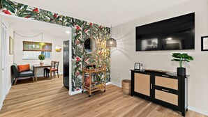 Una sala de estar moderna con piso de madera, una consola negra y de ratán, un televisor grande y un llamativo papel tapiz tropical.