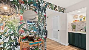 Una habitación con papel tapiz de temática tropical, un carrito de madera con diversos objetos, un espejo redondo y una zona de cocina con armarios verdes y platos decorativos.