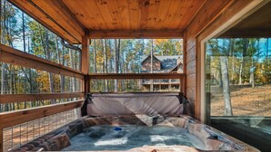A wooden cabin with a hot tub, large windows, and a view of a forested area. - LUXURY • Indoor Pool • Firepit • Suround sound Theater • Arcade • Sleeps 12 (Sevierville)