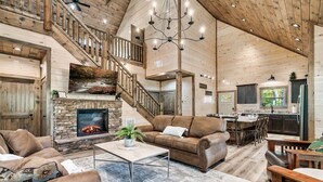 A spacious living room with a fireplace, leather sofas, a dining area, and a staircase leading to an upper level. - LUXURY • Indoor Pool • Firepit • Suround sound Theater • Arcade • Sleeps 12 (Sevierville)
