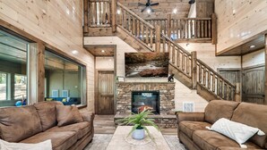 A spacious living room with a fireplace, wooden staircase, and two leather sofas. - LUXURY • Indoor Pool • Firepit • Suround sound Theater • Arcade • Sleeps 12 (Sevierville)