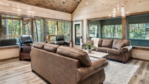 A spacious living room with a large brown leather sofa, a coffee table, and a fireplace. There are large windows offering views of a forested area. - LUXURY • Indoor Pool • Firepit • Suround sound Theater • Arcade • Sleeps 12 (Sevierville)