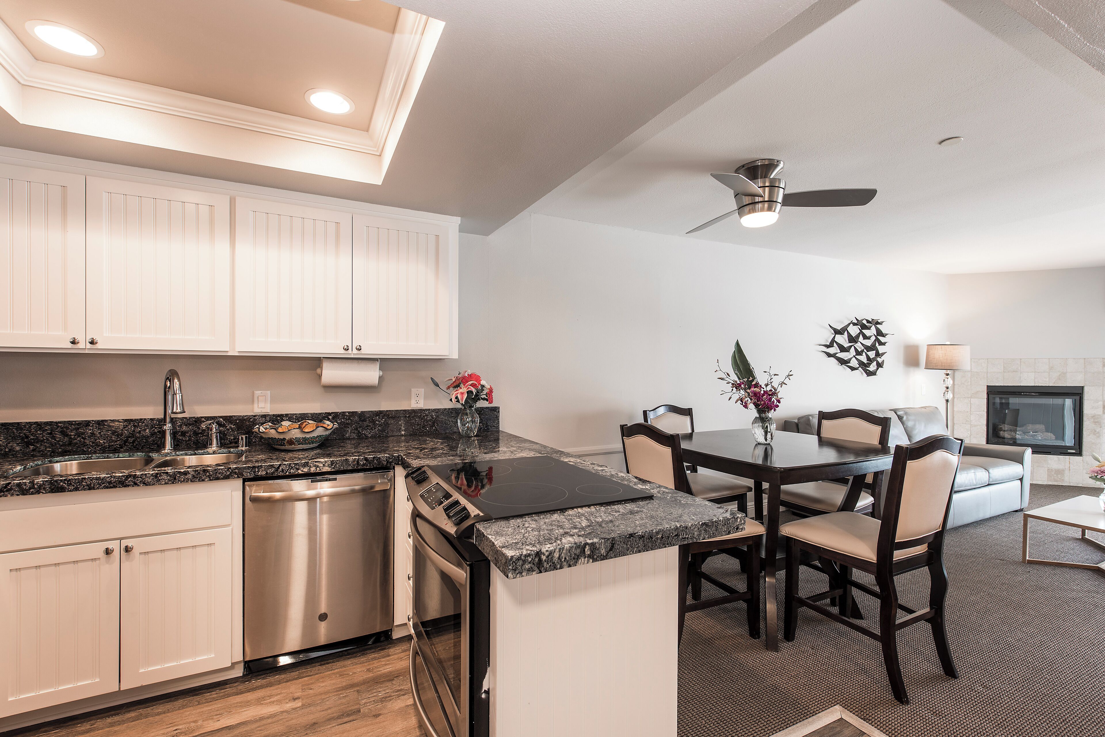 A modern kitchen with white cabinets, a built-in dishwasher, and a granite countertop. There is a dining area with a table and chairs, and a ceiling fan above.