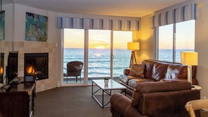 Un salon avec une cheminée, un canapé, une table basse et une vue sur l’océan au coucher du soleil.
