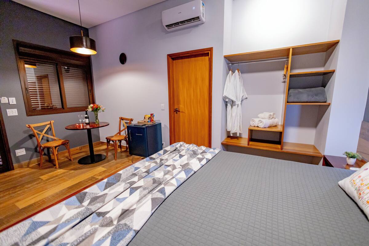 Habitación de hotel con cama, armario de madera, una pequeña mesa con sillas, escritorio y ventana con persianas.