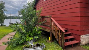 A red building with a wooden staircase leading to a deck, surrounded by greenery and a body of water in the background. - Birch Point Resort Kitchenette #14 - Lake Nelson, Hayward (Hayward)