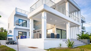 A modern two-story house with a balcony, large windows, and a covered outdoor seating area.