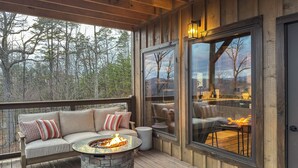 Una terraza de madera con una mesa de fuego de piedra, un área de descanso con cojines y grandes ventanales que ofrecen vistas a árboles y montañas.