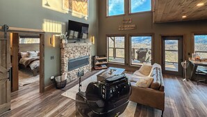 Amplia sala de estar con chimenea de piedra, ventanas grandes con vistas a las montañas, televisor de pantalla plana sobre la chimenea y una zona de estar acogedora con sofá de cuero y una mesita de centro.