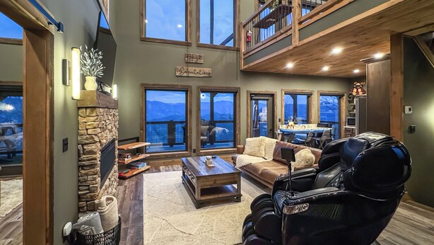 Un salón amplio con chimenea de piedra, ventanas grandes, techo alto y balcón en el segundo piso.