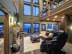 A spacious living room with a stone fireplace, large windows, a high ceiling, and a second-floor balcony.