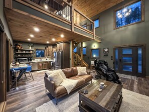 A spacious living area with a brown leather sofa, a coffee table, and a gaming chair. A kitchen with dark wooden cabinets and a refrigerator is visible in the background.