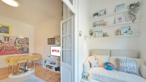A cozy living room with a sofa, a coffee table, and a TV. There are shelves with decorative items and a door leading to another room.
