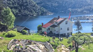 A white building with a red roof near a body of water, surrounded by greenery and hills.