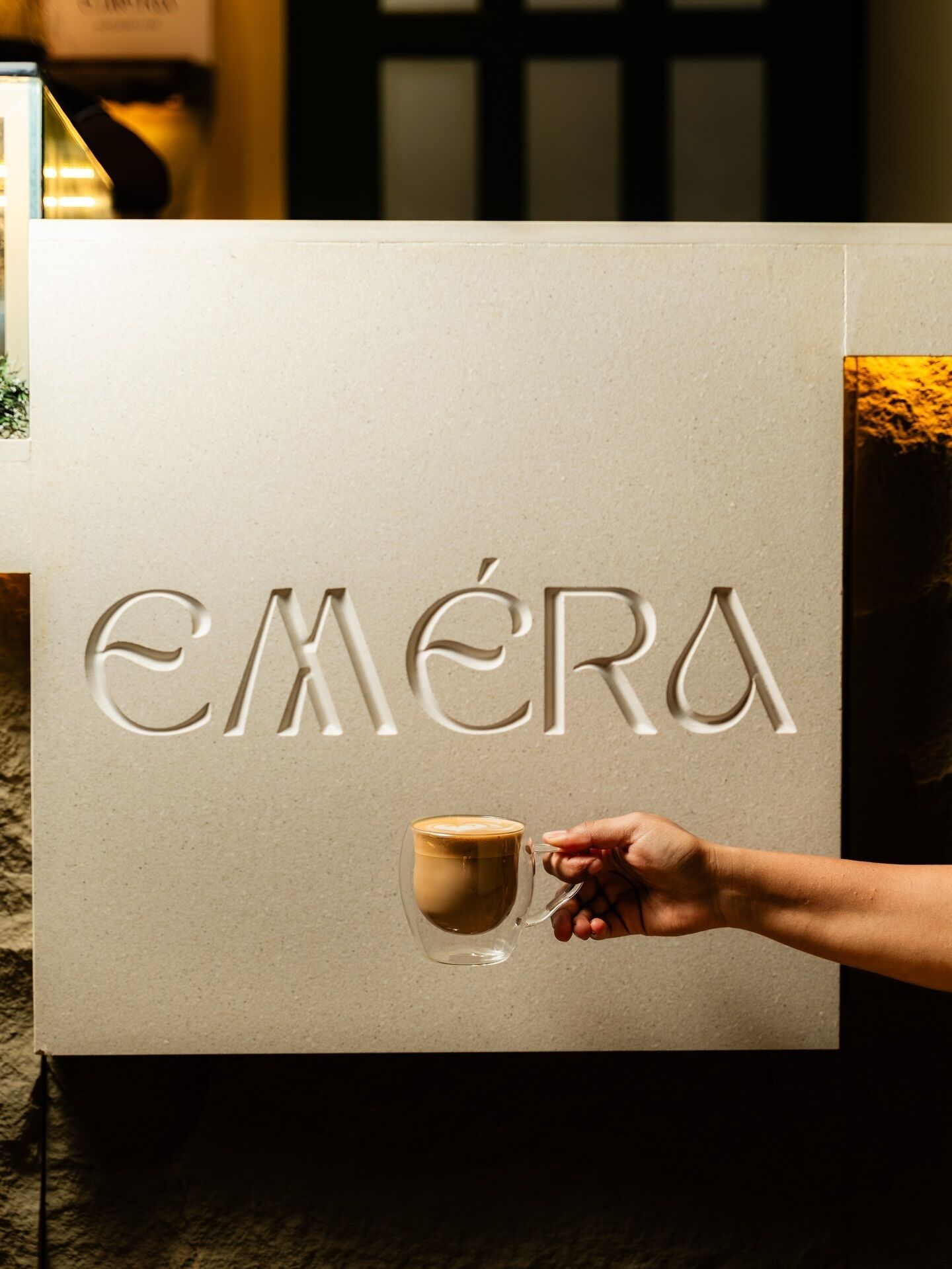 A hand holding a coffee cup in front of a sign with the word 'EMÉRA' embossed on it.