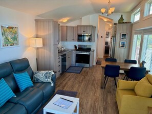 A modern kitchen with built-in appliances, a dining area with blue chairs, and a living room with a sofa and a yellow armchair.