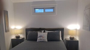 Un dormitorio con una cama grande, dos mesitas de noche con lámparas, una ventana pequeña sobre la cama y un cuadro enmarcado en la pared.