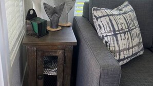 A corner of a room with a gray sofa, a patterned pillow, a small wooden side table with a lamp and decorative items, and a window with blinds. - 07 - Oceanside7 Cottage By The Sea at Alder Bay (Port McNeill)