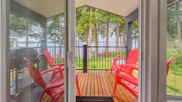 A covered deck with red chairs and a grill, overlooking a grassy area and trees. - 07 - Oceanside7 Cottage By The Sea at Alder Bay (Port McNeill)