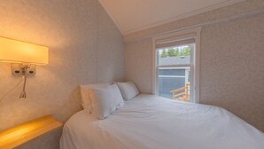 A bedroom with a bed, a nightstand, a wall-mounted lamp, and a window with a view of a wooden deck. - 07 - Oceanside7 Cottage By The Sea at Alder Bay (Port McNeill)