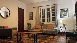 Ein Wohnzimmer mit Sofa, Couchtisch und Fernseher. Es gibt ein Fenster mit Vorhängen, einen Deckenventilator und einen Spiegel an der Wand.