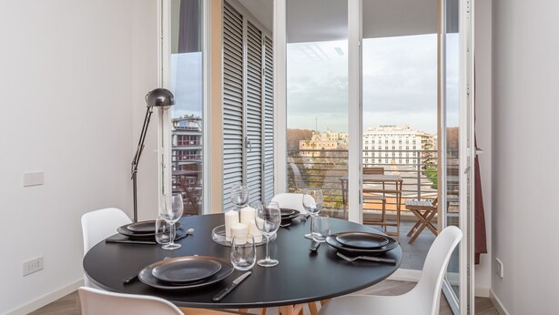 Ein Essbereich mit einem runden Tisch, gedeckt für vier Personen, einer schwarzen Tischdecke, weißen Stühlen und Blick auf Gebäude durch eine Glastür.