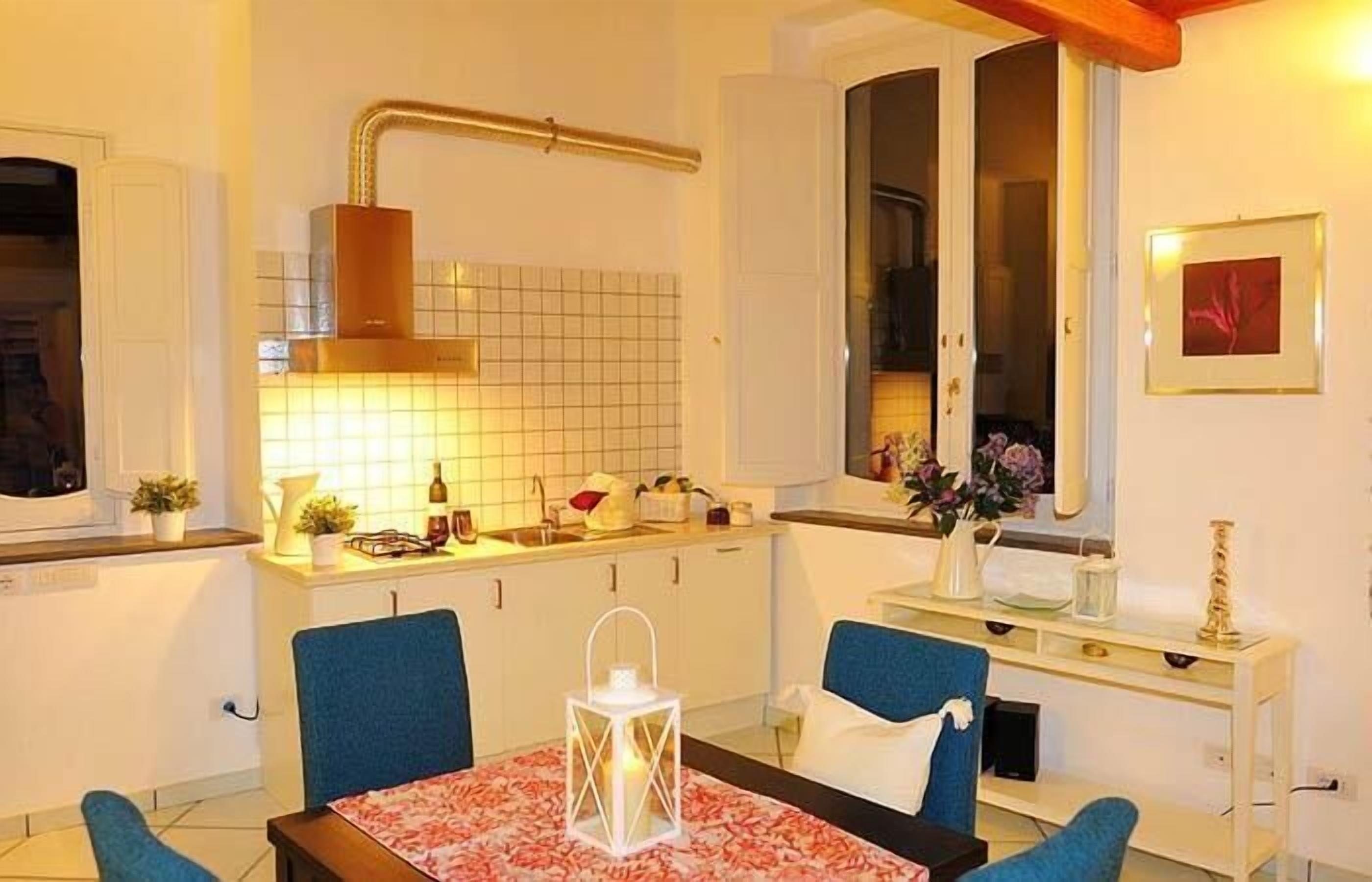 Une cuisine moderne avec des meubles blancs, un pare-feu en carrelage et un four encastré. On y trouve une table à manger avec des chaises bleues et une nappe blanche.
