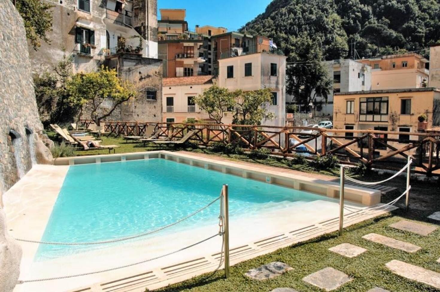 Una piscina con bordo in legno e poltrone prendisole, circondata da edifici con muri in pietra e un paesaggio montuoso alle spalle.